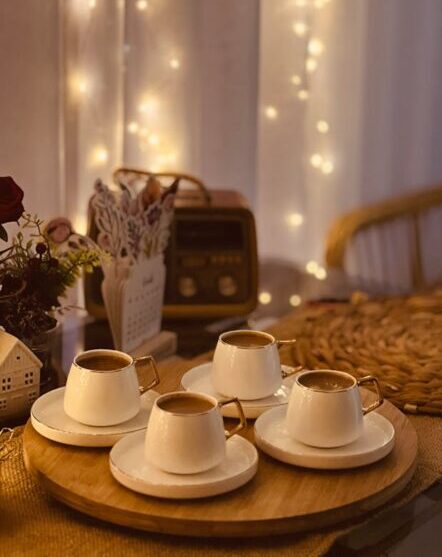 A warm indoor scene with four cups of coffee on a wooden tray, surrounded by cozy decor and fairy lights.