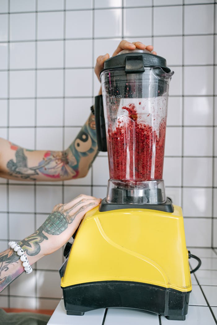 Tattooed hands using a yellow blender to mix red fruits in a tiled kitchen setting.